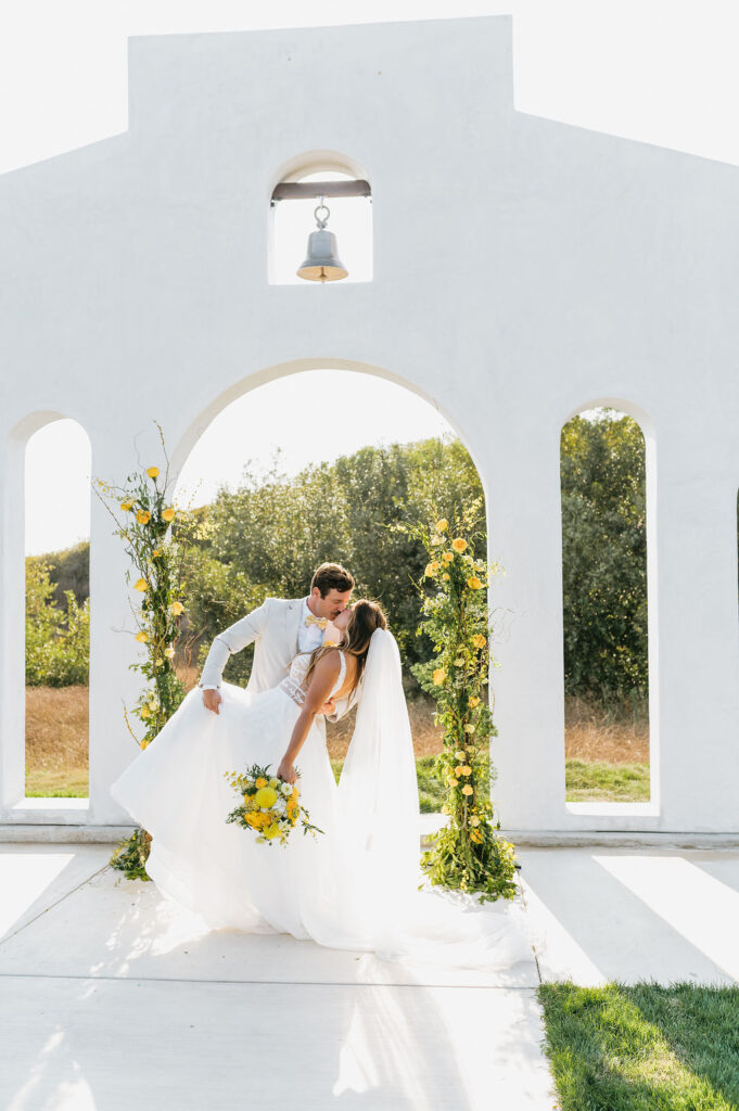 Bride in white dress and groom in tan suit kiss in front of the ceremony site arch backdrop at Jeune Perche Wedding Estate