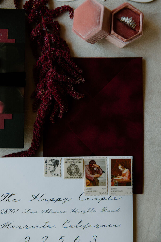 White envelope with black script calligraphy and vintage postage stamps, laying on top of deep burgundy velvet envelope. Vintage engagement ring in blush velvet ring box and burgundy flower.