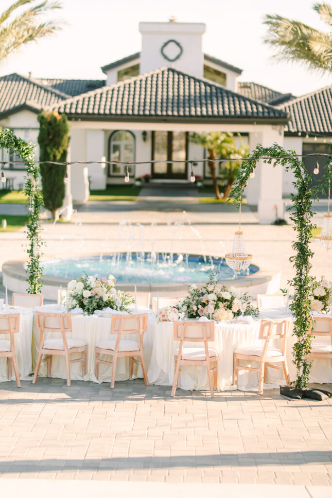 Outdoor wedding reception setup at The Hillside Estate private estate wedding venue in San Marcos, California, with round guest tables, white linens, and floral centerpieces surrounding the estate fountain with string lights and the main house in the background.