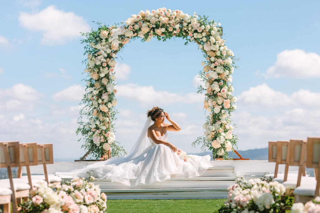 Bride seated on ceremony stage beneath a floral arch with panoramic sky and hillside views at The Hillside Estate private estate wedding venue in San Marcos, California.
