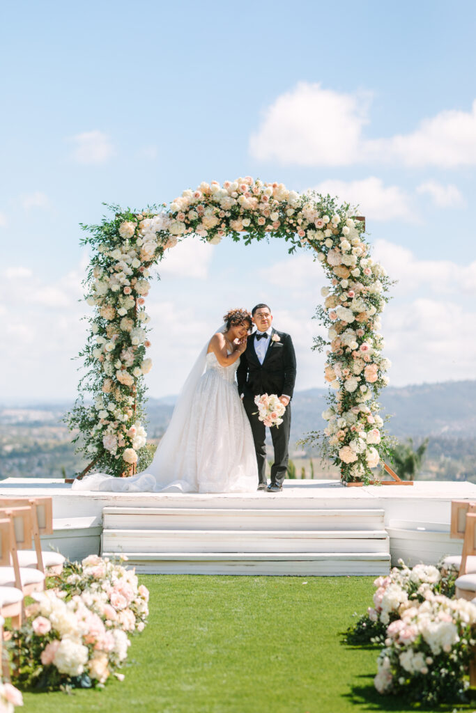 Bride and groom standing together on the ceremony stage beneath a floral arch with rolling valley views at The Hillside Estate.