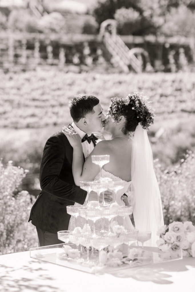 Black and white image of bride and groom kissing behind a champagne tower during cocktail hour at The Hillside Estate.