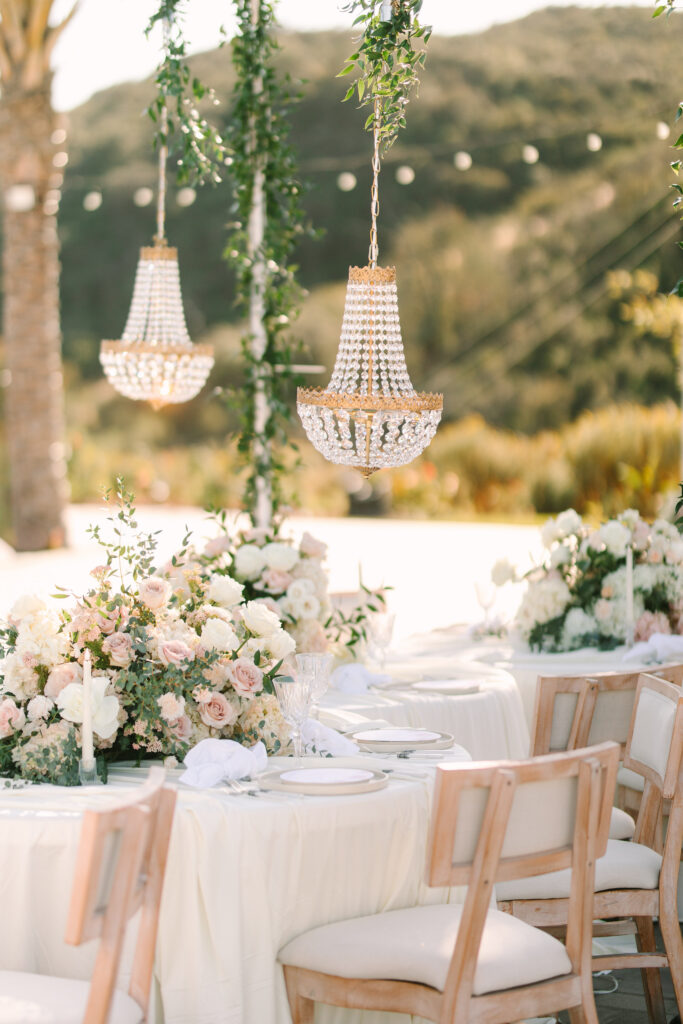 Crystal chandeliers hanging over floral-lined reception tables with string lights and hillside views at The Hillside Estate private estate wedding venue in San Marcos, California.