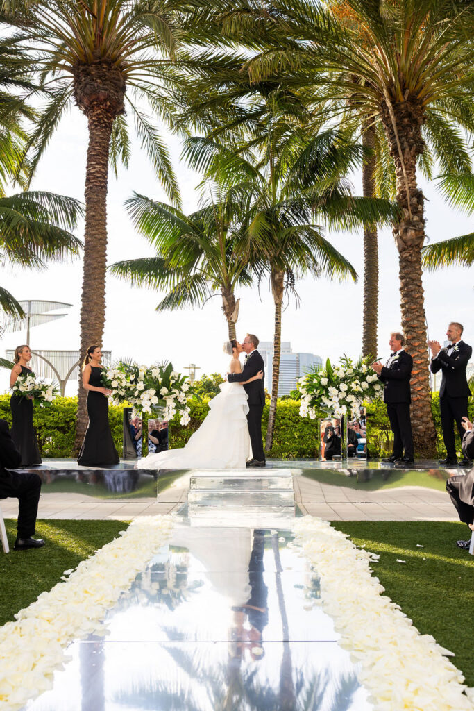 Bride and groom kiss at the altar on a mirrored stage flanked by the bridal party in black attire, white floral arrangements, and palm trees at a Hilton San Diego Bayfront wedding