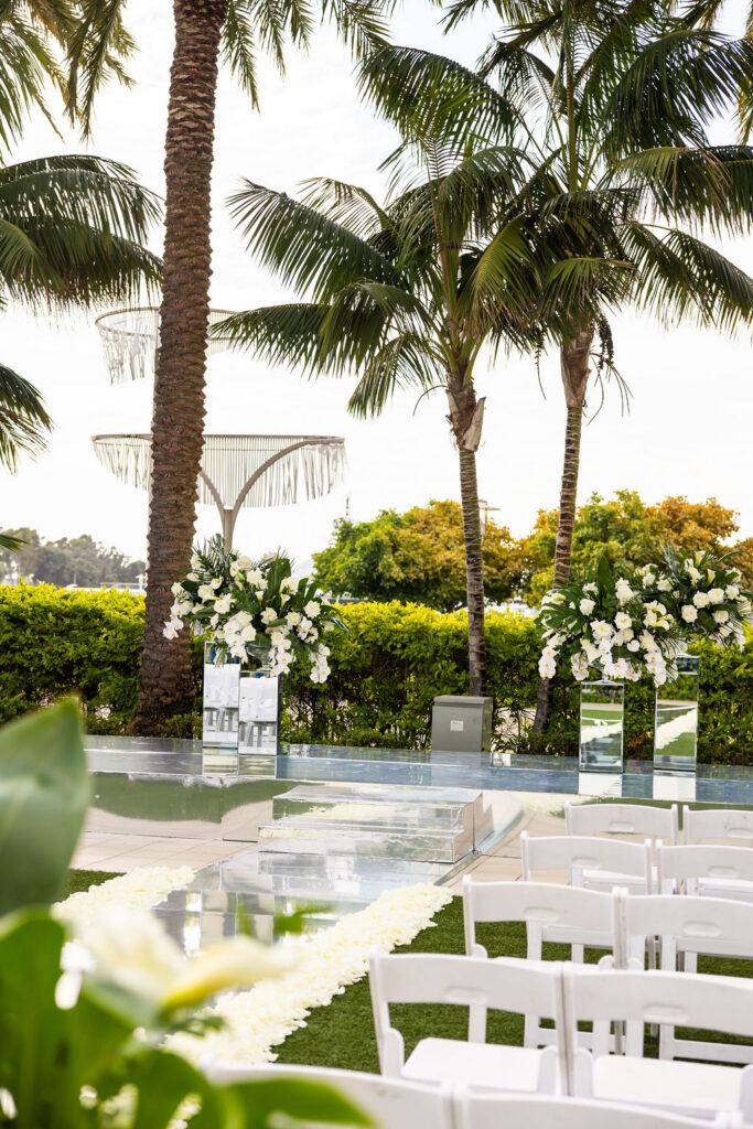 Close up of the mirrored wedding ceremony stage altar at the Hilton San Diego Bayfront, flanked by lush white floral arrangements with orchids and tropical greenery, with a rose petal lined mirror aisle in the foreground