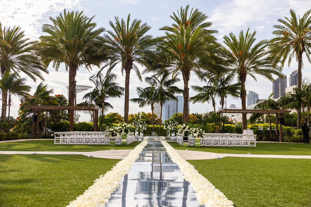 Ground level view looking down the mirrored aisle runner at a Hilton San Diego Bayfront wedding ceremony setup, framed by palm trees and the San Diego skyline with white chairs and white floral arrangements on either side