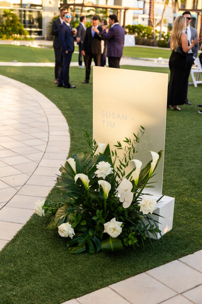 Modern white wedding welcome sign reading Susan and Tim on frosted acrylic surrounded by white calla lilies, roses, and tropical greenery at the Hilton San Diego Bayfront