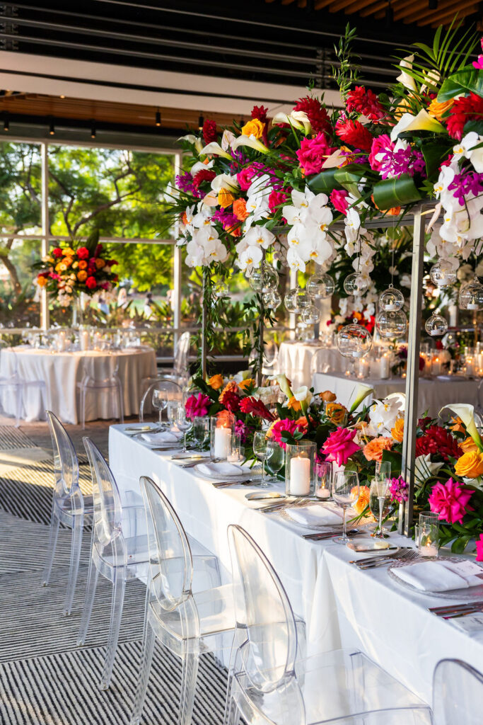 Tropical wedding reception tablescape with vibrant pink and orange florals, white orchids, hanging glass globes, and ghost chairs at a Hilton San Diego Bayfront wedding
