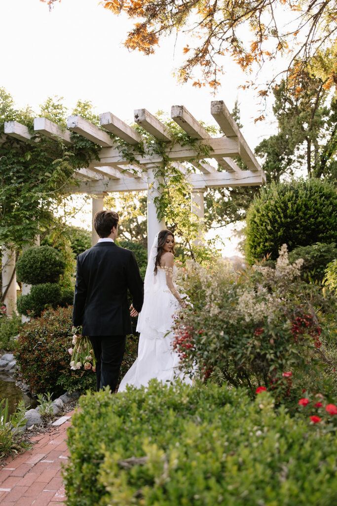 Bride and groom walking along a brick pathway toward a pergola surrounded by lush gardens at golden hour at this Temecula wedding venue.